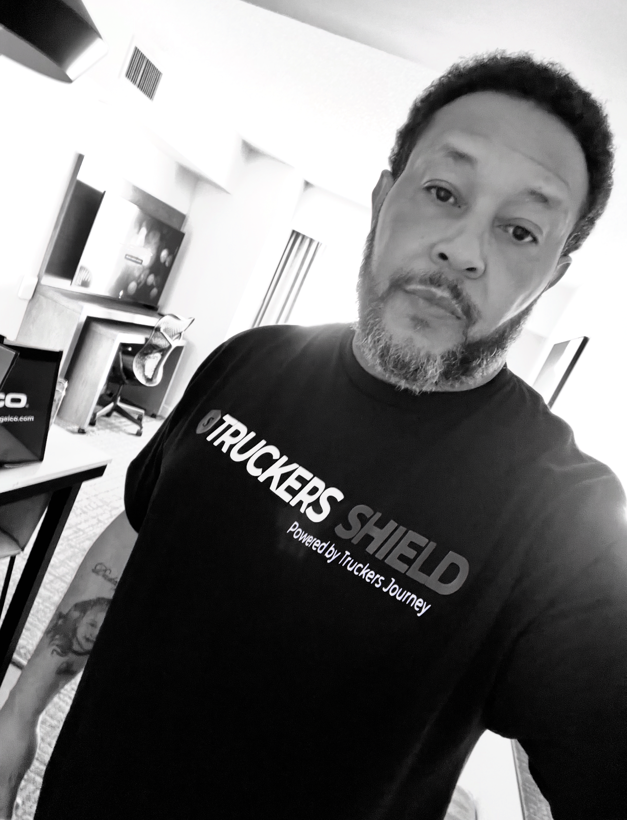 Candid black-and-white portrait of Goins Digital founder Darius Goins wearing a Truckers Shield t-shirt, with a visible forearm tattoo, photographed indoors